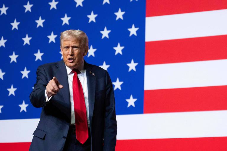 Donald Trump veste terno azul escuro, camisa branca e gravata vermelha, apontando com a mão direita enquanto fala. Ao fundo, grande bandeira dos Estados Unidos com estrelas brancas em campo azul e listras vermelhas e brancas.