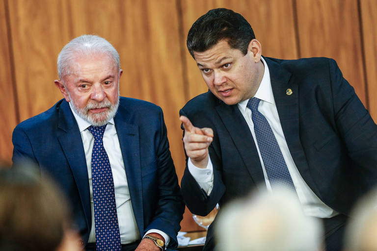 Dois homens sentados lado a lado em ambiente com parede de madeira ao fundo. O homem à esquerda tem cabelo e barba grisalhos, veste terno azul escuro, camisa branca e gravata azul com padrão. O homem à direita, de cabelo escuro, veste terno preto, camisa branca e gravata azul, e aponta com o dedo indicador enquanto fala.