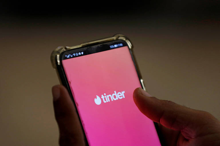 Mão segurando smartphone com a tela exibindo o logo do aplicativo Tinder em fundo degradê rosa e laranja.