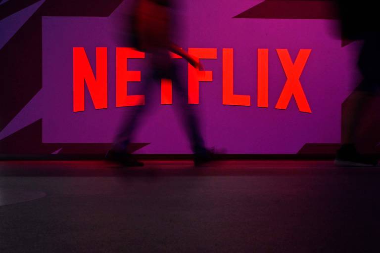 Logo vermelho da Netflix iluminado em fundo roxo com silhuetas desfocadas de pessoas caminhando em frente.
