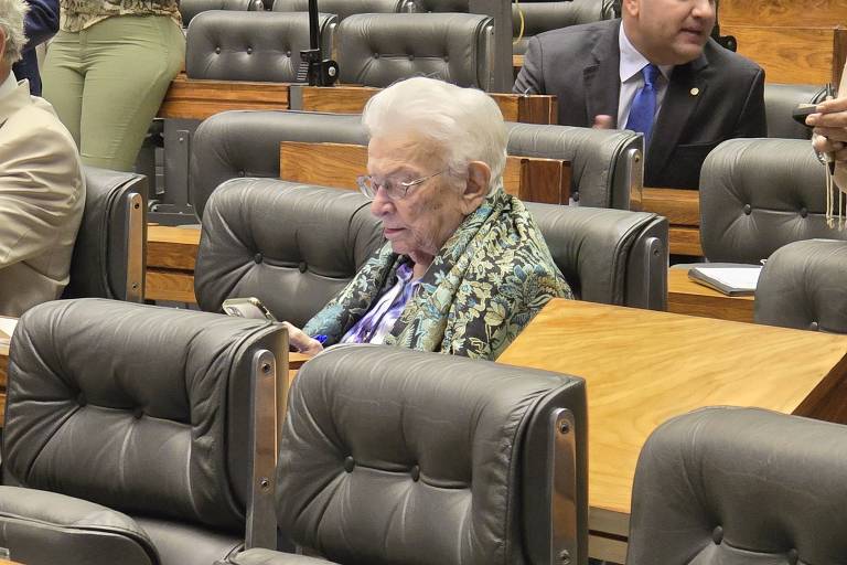Mulher idosa com cabelo branco e cachecol colorido sentada em cadeira de couro marrom em plenário com várias cadeiras vazias ao redor. Pessoas ao fundo em trajes formais.
