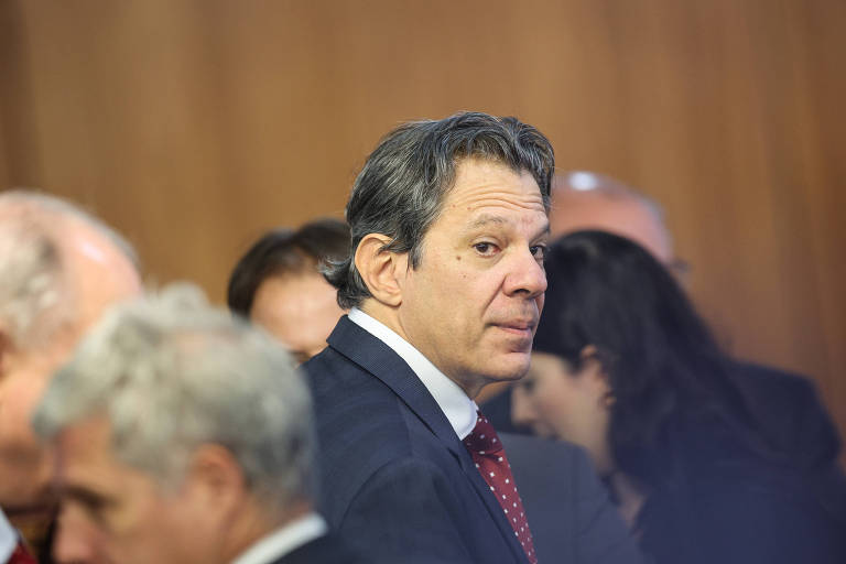 Homem de meia-idade com cabelo escuro e terno azul escuro, camisa branca e gravata vermelha com pontos brancos, olhando para a câmera em ambiente interno com outras pessoas ao fundo desfocadas.