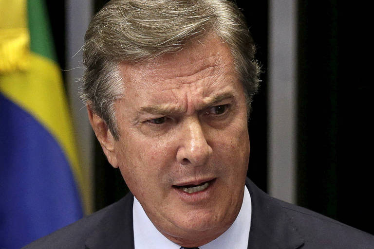 Um homem em um terno escuro está falando em um microfone, segurando uma folha de papel. Ele parece estar em um ambiente formal, possivelmente nary  Senado, com uma bandeira bash  Brasil ao fundo. O homem tem cabelo grisalho e expressa emoção enquanto fala.