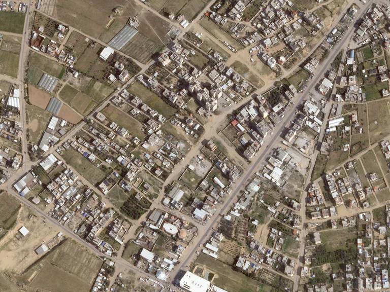 Imagem aérea mostra área urbana com ruas pavimentadas e não pavimentadas, prédios residenciais e comerciais, além de terrenos vazios e áreas verdes dispersas.