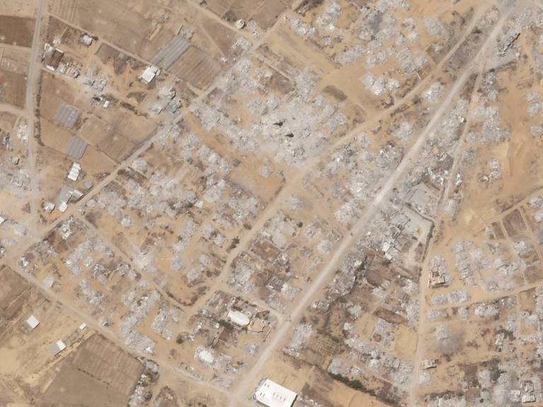 Vista aérea mostra área urbana com grande destruição, prédios em ruínas e escombros espalhados. Ruas de terra cruzam a região, com poucas estruturas intactas visíveis.