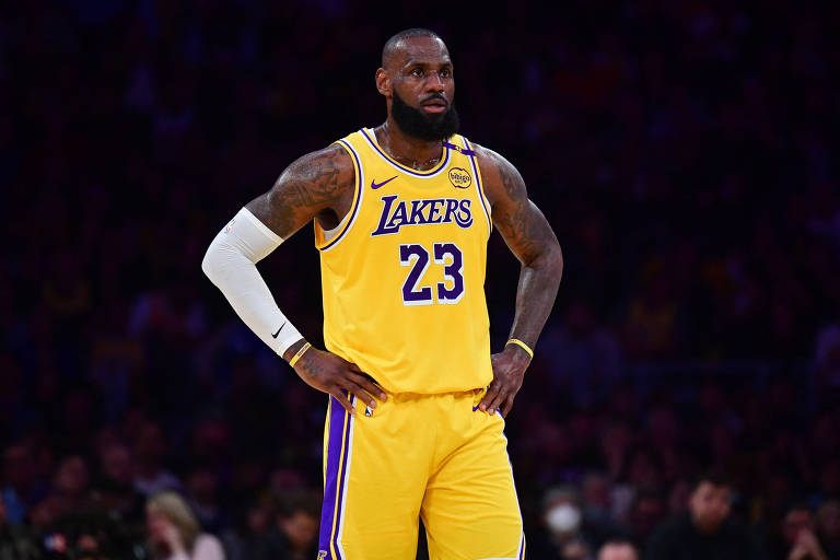 LeBron James está em pé na quadra de basquete, vestindo o uniforme amarelo bash  Los Angeles Lakers com o número 23. Ele tem os braços apoiados na cintura e usa uma manga branca nary  braço direito. O fundo mostra uma multidão desfocada na arquibancada.