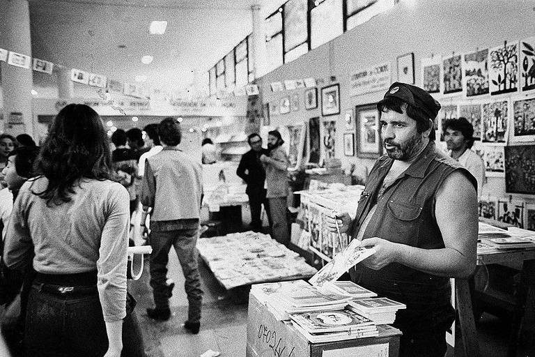 Homem com colete sem mangas e boina segura uma publicação em feira interna de livros e zines. Pessoas circulam entre mesas e paredes decoradas com cartazes e ilustrações.
