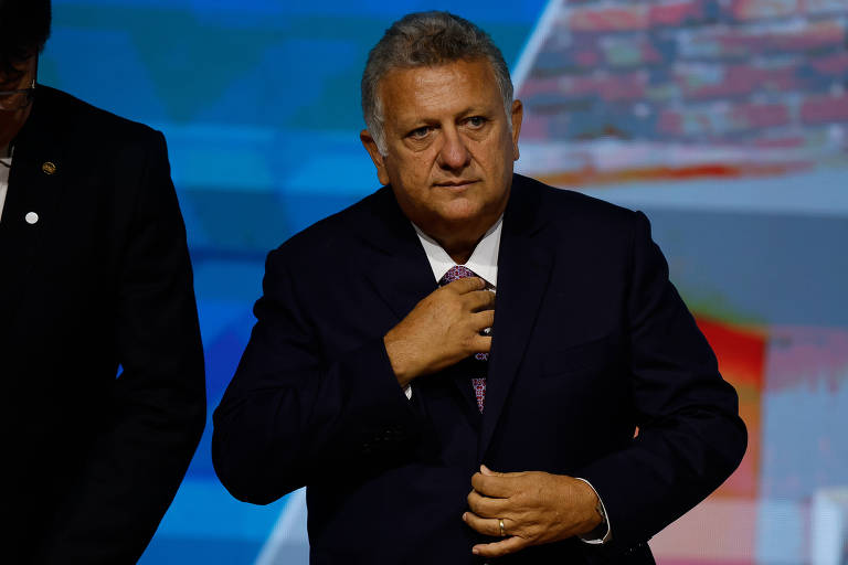 Homem de cabelos grisalhos veste terno escuro e ajusta a gravata em ambiente com fundo desfocado que sugere palco ou evento