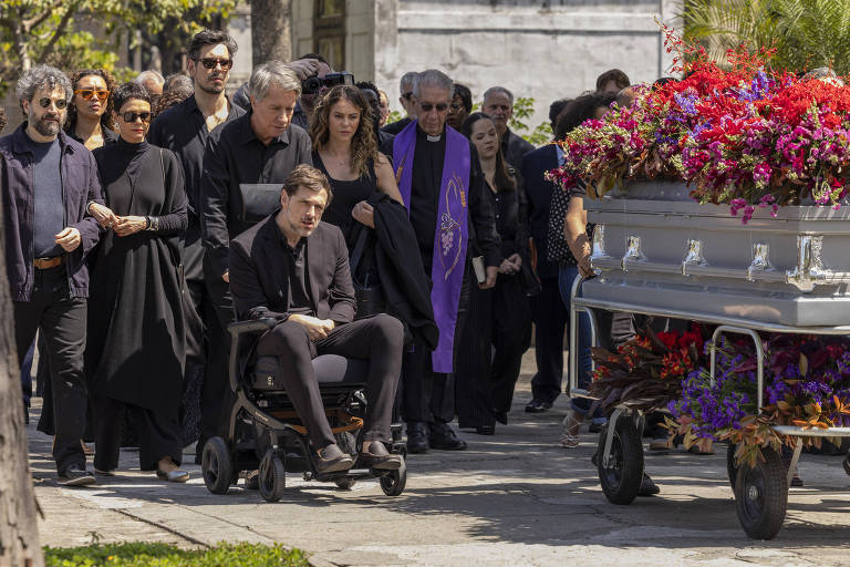 Grupo de pessoas vestidas de preto acompanha caixão cinza decorado com flores vermelhas e roxas em carrinho. Homem em cadeira de rodas está à frente, seguido por sacerdote com estola roxa e demais presentes em ambiente externo.