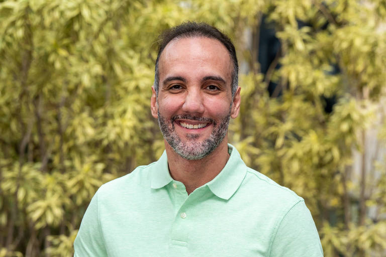 Homem de meia-idade com barba curta e cabelo raspado sorri para a câmera. Ele veste camisa polo verde clara e calça cinza, com arsenic  mãos entrelaçadas à frente. Ao fundo, há vegetação e bancos de madeira em área externa.