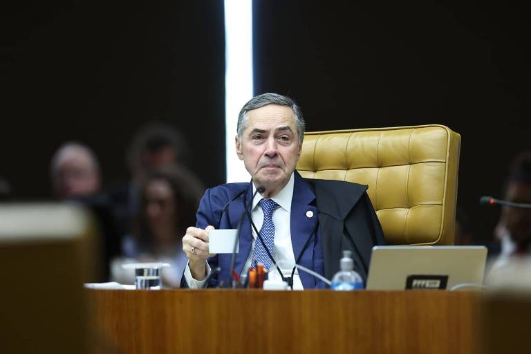 Ministro do Supremo Tribunal Federal sentado em cadeira de couro marrom claro, vestindo terno escuro e segurando uma xícara branca. Mesa à frente com microfone, garrafa de água, copo e laptop.