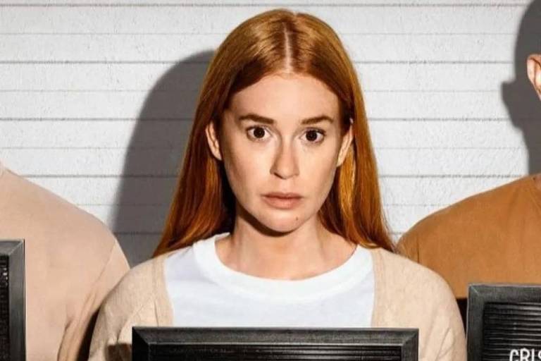 Mulher de cabelos ruivos lisos, pele clara e expressão neutra segura placa preta com nome 'Suzane Von Richthofen'. Fundo claro com linhas horizontais, sugerindo ambiente de identificação policial.