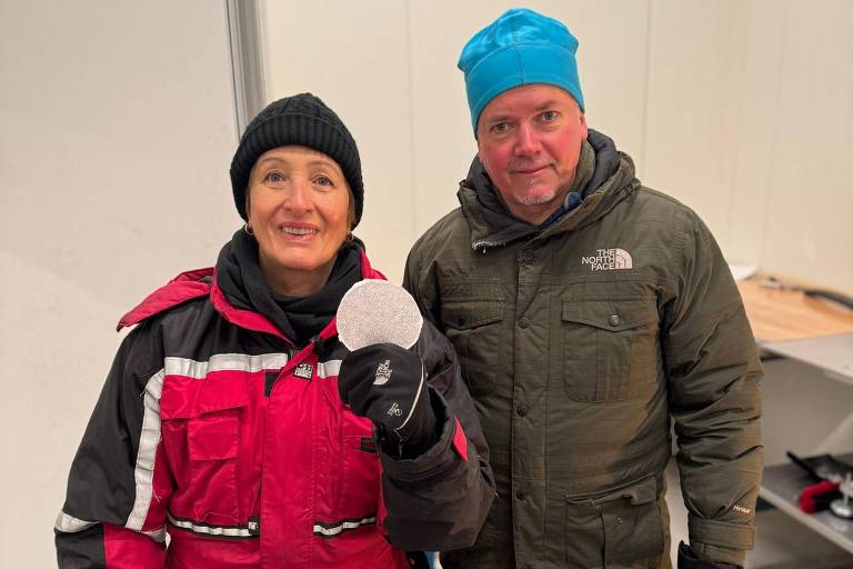 Duas pessoas posam em ambiente interno com paredes brancas. A pessoa à esquerda veste macacão vermelho com detalhes pretos e brancos, gorro preto e luvas pretas, segurando um objeto redondo branco. A pessoa à direita usa casaco verde escuro, gorro azul e luvas pretas.