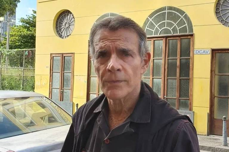 Em foto colorida, homem de camisa preta tira foto em frente a um prédio hiatórico