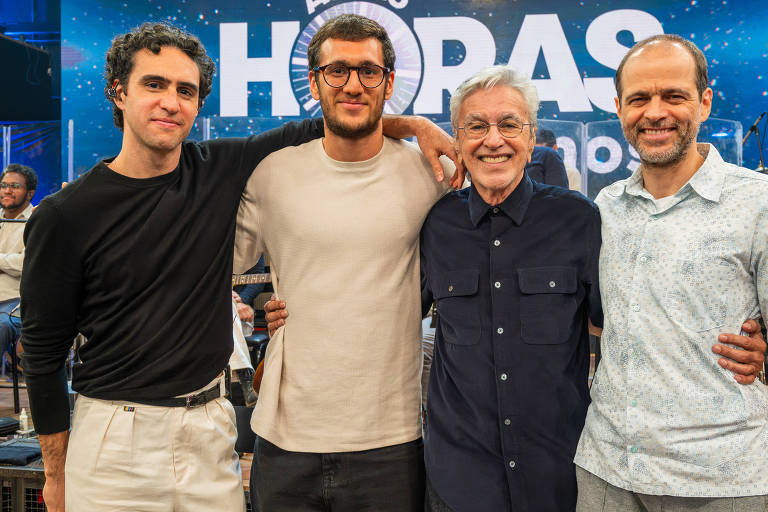 Quatro homens estão lado a lado, com os braços nos ombros uns dos outros, sorrindo para a câmera em estúdio iluminado. Ao fundo, painel azul com texto 'Altas Horas'. Eles vestem roupas casuais, e há equipamentos de gravação e outras pessoas sentadas ao fundo.