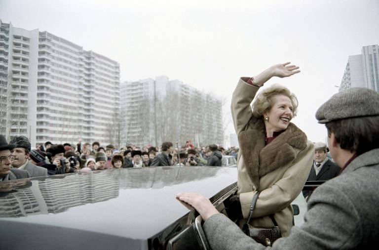 A woman in a beige coat with a fur collar waves and smiles to a crowd gathered in an urban area with tall residential buildings in the background. A man in a gray hat stands beside her, holding the door of his black car.