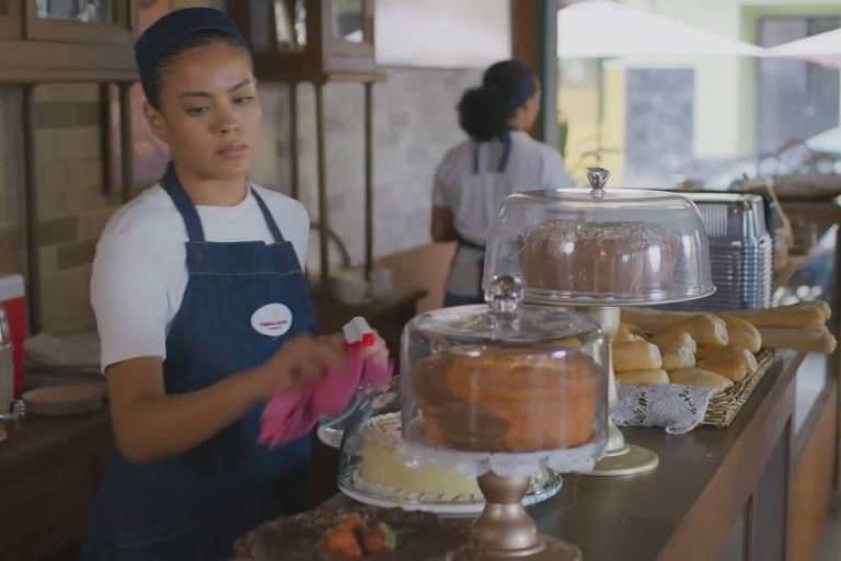 Mulher com avental azul e camiseta branca limpa a vitrine de bolos em balcão de confeitaria. Ao fundo, outra funcionária e mesas externas visíveis.