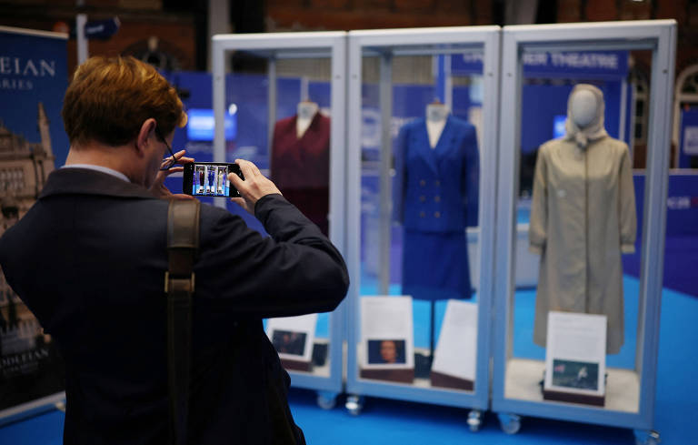 A man with his back turned takes a cell phone photo of three mannequins displaying vintage women's clothing in glass display cases. The pieces include a blue dress, a beige coat, and a burgundy suit, each accompanied by informational signs.