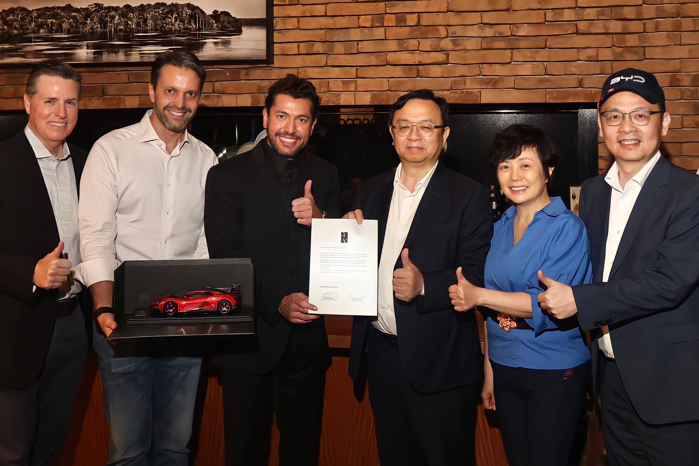 Seis pessoas em ambiente interno com parede de tijolos ao fundo posam para foto. Dois homens no centro seguram um documento e uma caixa com miniatura de carro vermelho. Todos fazem sinal de positivo com o polegar.