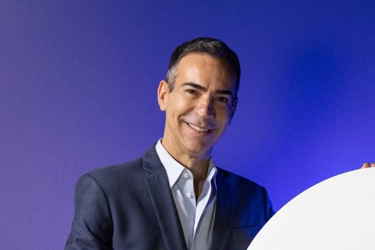 Homem vestido com terno azul escuro e camisa branca está ao lado de um grande logotipo circular da Globo, apoiado sobre um pedestal azul escuro, em fundo azul.