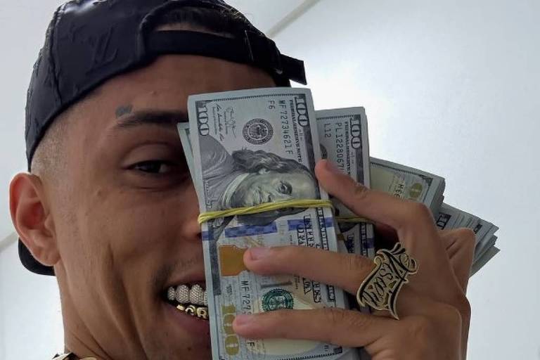 Homem com boné preto e dentes com grill segura maço de dólares próximo ao rosto. Ele usa colares dourados grandes, anel e relógio prateado, e tem tatuagens no peito.