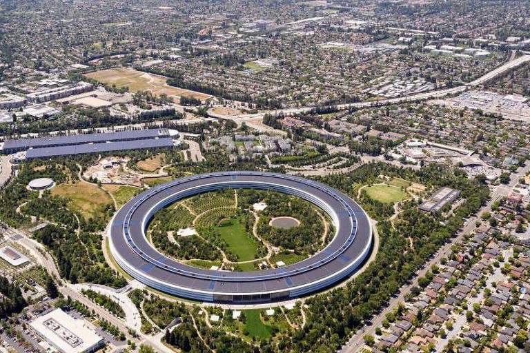 Campus circular da Apple em Cupertino cercado por áreas verdes e edifícios. Residências e ruas organizadas em grade ocupam a área ao redor, com montanhas ao fundo sob céu claro.