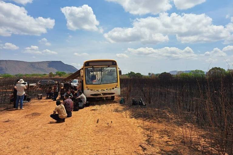 Ônibus escolar amarelo parado em estrada de terra em área rural. Pessoas sentadas e agachadas próximas ao ônibus. Vegetação ao redor está queimada, com galhos secos e pretos. Céu azul com nuvens dispersas e montanhas ao fundo.