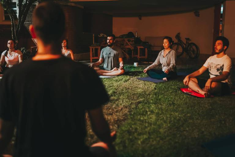 Seis pessoas sentadas em círculo no gramado, com as pernas cruzadas e olhos fechados, praticando meditação. Um instrutor está de costas para a câmera, conduzindo a sessão em ambiente externo com iluminação suave.