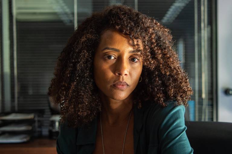 Mulher de cabelos cacheados e escuros está sentada em cadeira preta atrás de mesa de vidro em escritório. Ao fundo, há estantes com papéis e janela com persianas fechadas.