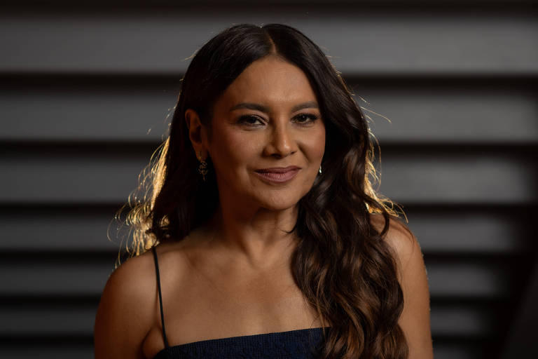 Mulher com cabelo longo e ondulado, usando vestido de alças finas azul escuro, posando em fundo desfocado com linhas horizontais escuras.