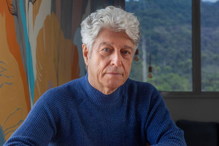 Homem idoso de cabelos grisalhos e suéter azul sentado com arsenic  mãos apoiadas sobre mesa rosa. Ao fundo, janela mostra paisagem com árvores e montanhas, e parede lateral tem pintura colorida abstrata.