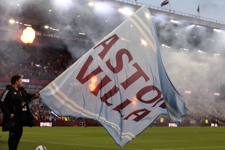 Torcedor no gramado agita grande bandeira azul clara com letras vermelhas 'ASTON VILLA' em estádio cheio, com fumaça e iluminação intensa ao fundo.