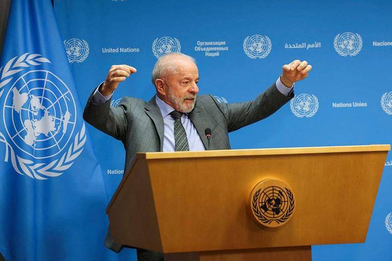 Homem de terno cinza e gravata verde fala em púlpito da ONU, gesticulando com os braços levantados. Fundo azul exibe logotipos da ONU e bandeira da organização à esquerda.