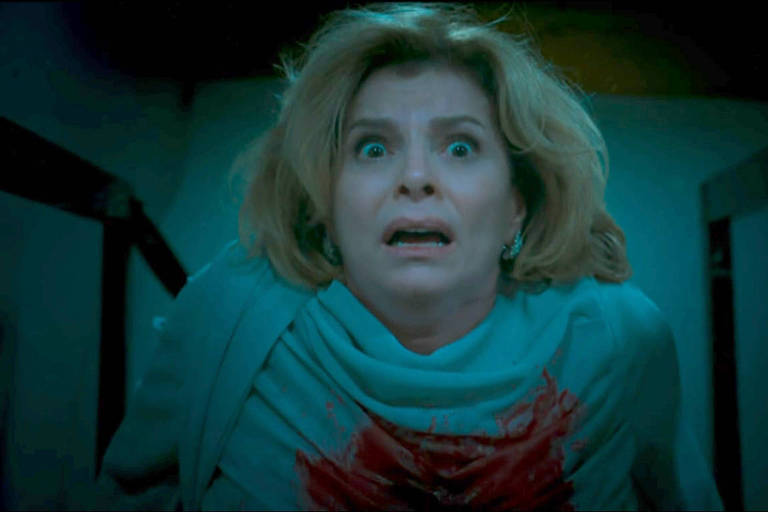 Mulher de cabelos curtos e loiros veste blusa azul com mancha vermelha que parece sangue nary  peito. Ela olha para frente com olhos arregalados e boca entreaberta, em ambiente escuro.