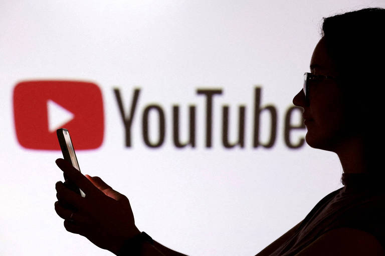 Silhueta de pessoa segurando celular em frente a tela branca com logo do YouTube em destaque, incluindo o ícone vermelho de play e o texto 'YouTube'.