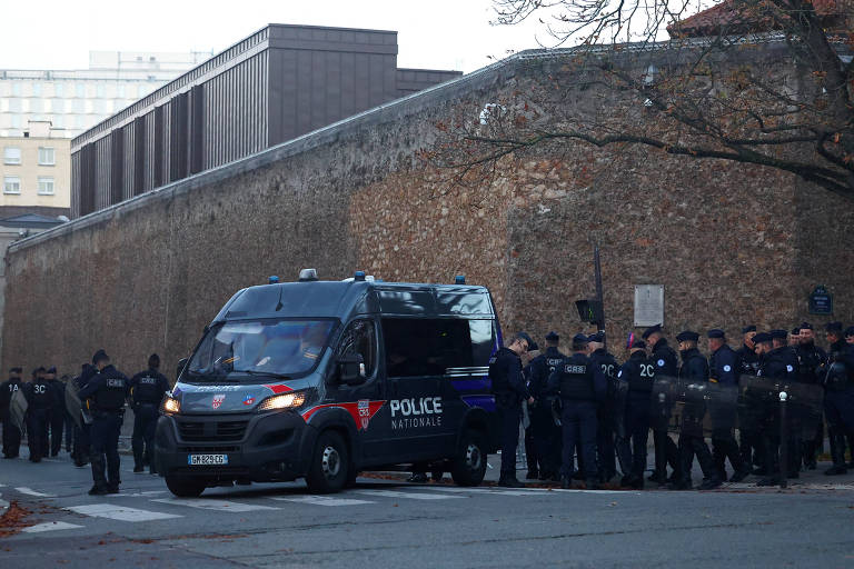 Grupo de policiais nacionais em uniforme preto reunidos ao lado de uma van preta da polícia, estacionada próxima a um muro alto de pedra em área urbana durante o dia.
