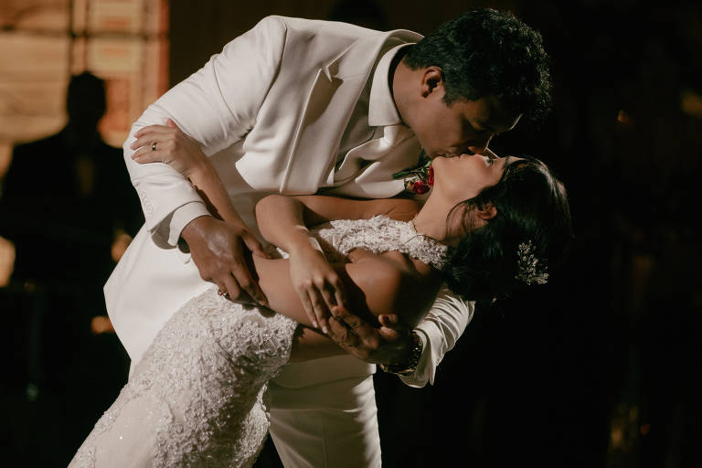 Noivo vestido com terno branco inclina a noiva em vestido branco rendado para um beijo durante a dança. Fundo escuro destaca o casal em primeiro plano.