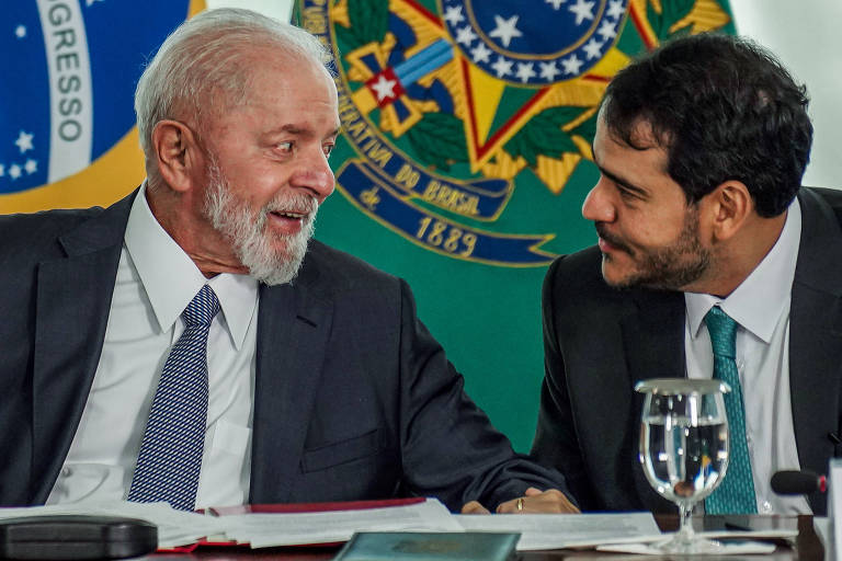 Dois homens de terno, um mais velho com cabelo e barba grisalhos e outro mais jovem com cabelo escuro, sentados lado a lado em mesa com documentos e copo de água. Ao fundo, brasão da República Federativa do Brasil em destaque.