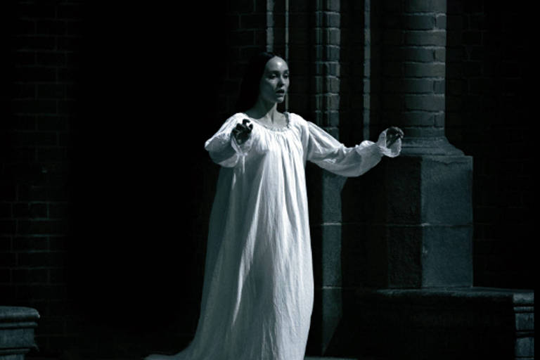 A imagem mostra uma figura feminina vestida com um longo vestido branco, que parece estar em uma escadaria. A cena é iluminada de forma sombria, com um fundo escuro e paredes de tijolos. A mulher está com os braços estendidos, como se estivesse chamando ou se movendo lentamente. A atmosfera é de mistério e suspense, evocando uma sensação de assombração.