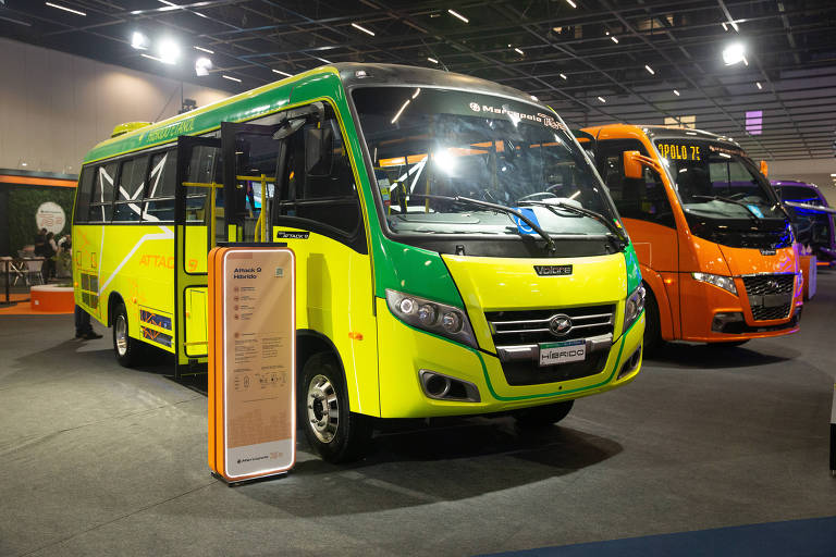 Micro-ônibus verde e amarelo com portas abertas em salão de exposição. Ao fundo, outro micro-ônibus laranja está estacionado sob iluminação artificial.