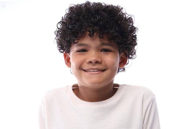 Menino de cabelos cacheados e escuros usa camiseta branca e segura um volante preto, posicionado contra fundo branco.
