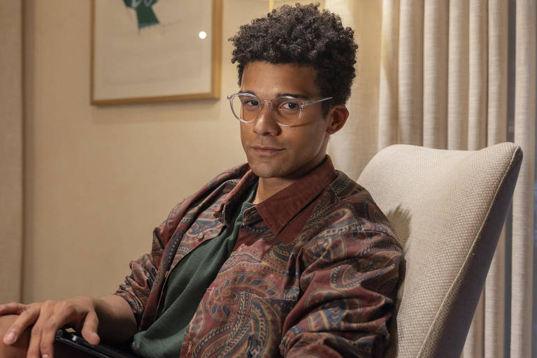 Um jovem sentado em uma cadeira confortável, usando óculos e uma camisa de manga curta verde sob uma jaqueta estampada. Ele tem cabelo cacheado e está olhando para a câmera com uma expressão neutra. Ao fundo, há cortinas claras e uma parede com uma obra de arte emoldurada.