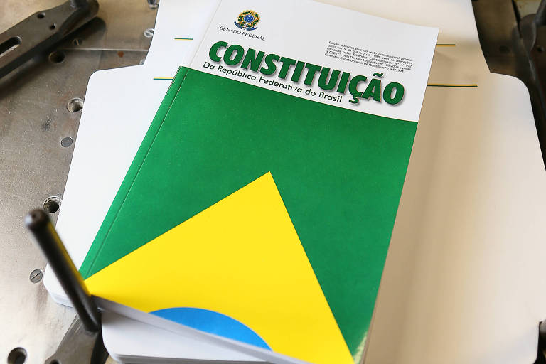 Livro da Constituição da República Federativa do Brasil com capa verde, amarela e azul, posicionado sobre uma máquina de impressão metálica com alavancas e parafusos visíveis.