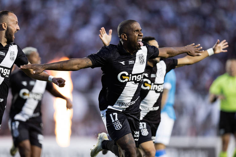 Três jogadores do time com uniforme preto e branco comemoram com os braços abertos durante partida de futebol. O jogador central, com número 17, está em destaque, expressando entusiasmo. Ao fundo, público desfocado e árbitro com uniforme verde.