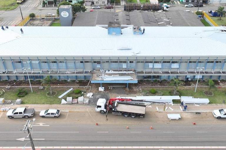 Prédio comercial retangular de dois andares com fachada azul clara e telhado branco, localizado em área urbana com ruas e casas ao redor. Na frente do prédio, há cinco veículos estacionados, incluindo um caminhão com guindaste em operação. A rua em primeiro plano tem faixa de pedestres e postes de energia.