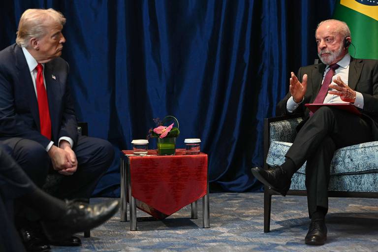 Donald Trump e Luiz Inácio Lula da Silva sentados em poltronas opostas com mesa pequena entre eles, contendo dois copos e um vaso com flor. Fundo com cortina azul e parte da bandeira do Brasil visível.