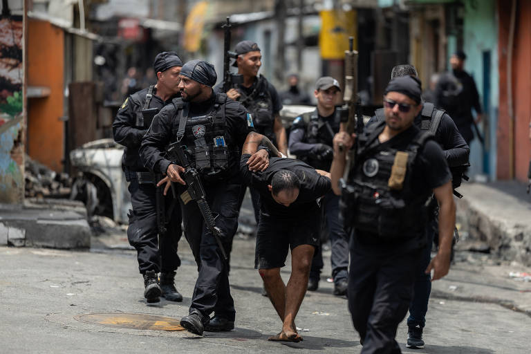 Seis policiais militares armados escoltam homem detido, que está curvado e com as mãos para trás, em rua estreita de comunidade com casas coloridas e paredes desgastadas.