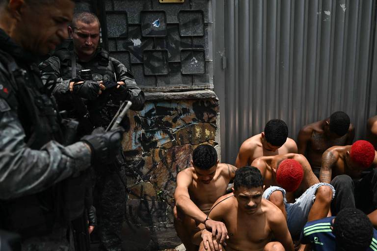 Dois policiais armados e uniformizados observam seis homens agachados, sem camisa, com arsenic  mãos para trás, encostados em parede grafitada e portão metálico.