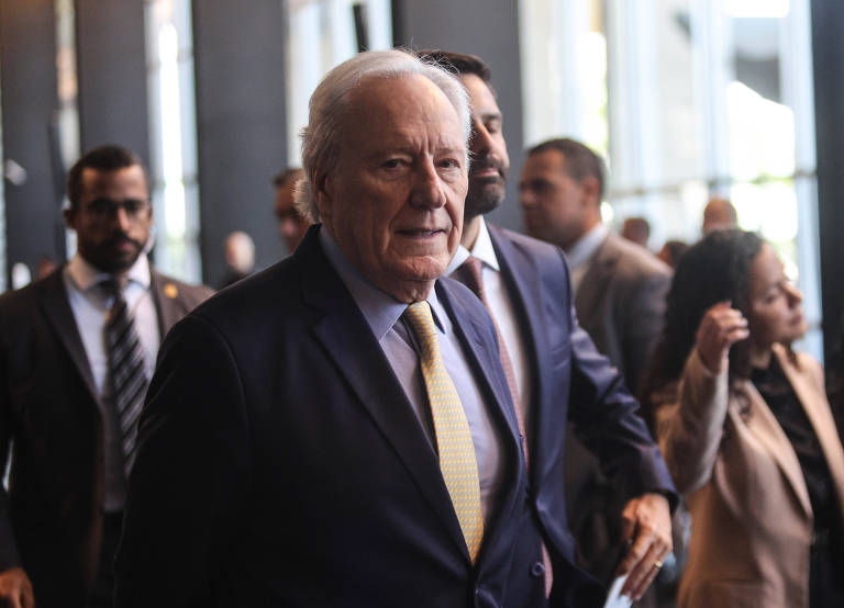 Homem idoso de cabelo branco veste terno escuro, camisa azul clara e gravata amarela, caminhando em corredor interno com outras pessoas ao fundo, algumas em trajes formais. Ambiente iluminado por luz natural de janelas grandes.