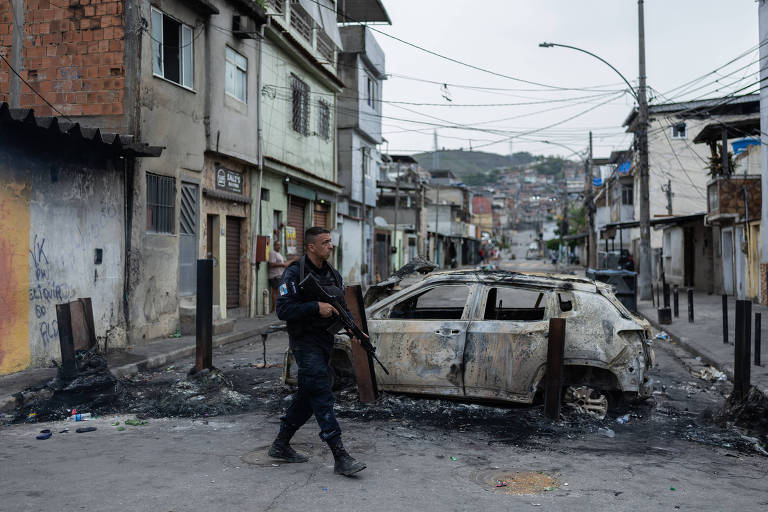 Policial com fuzil caminha em rua de bairro com casas simples e carro completamente queimado bloqueando a via. Resquícios de incêndio e sujeira no chão indicam recente conflito ou vandalismo.